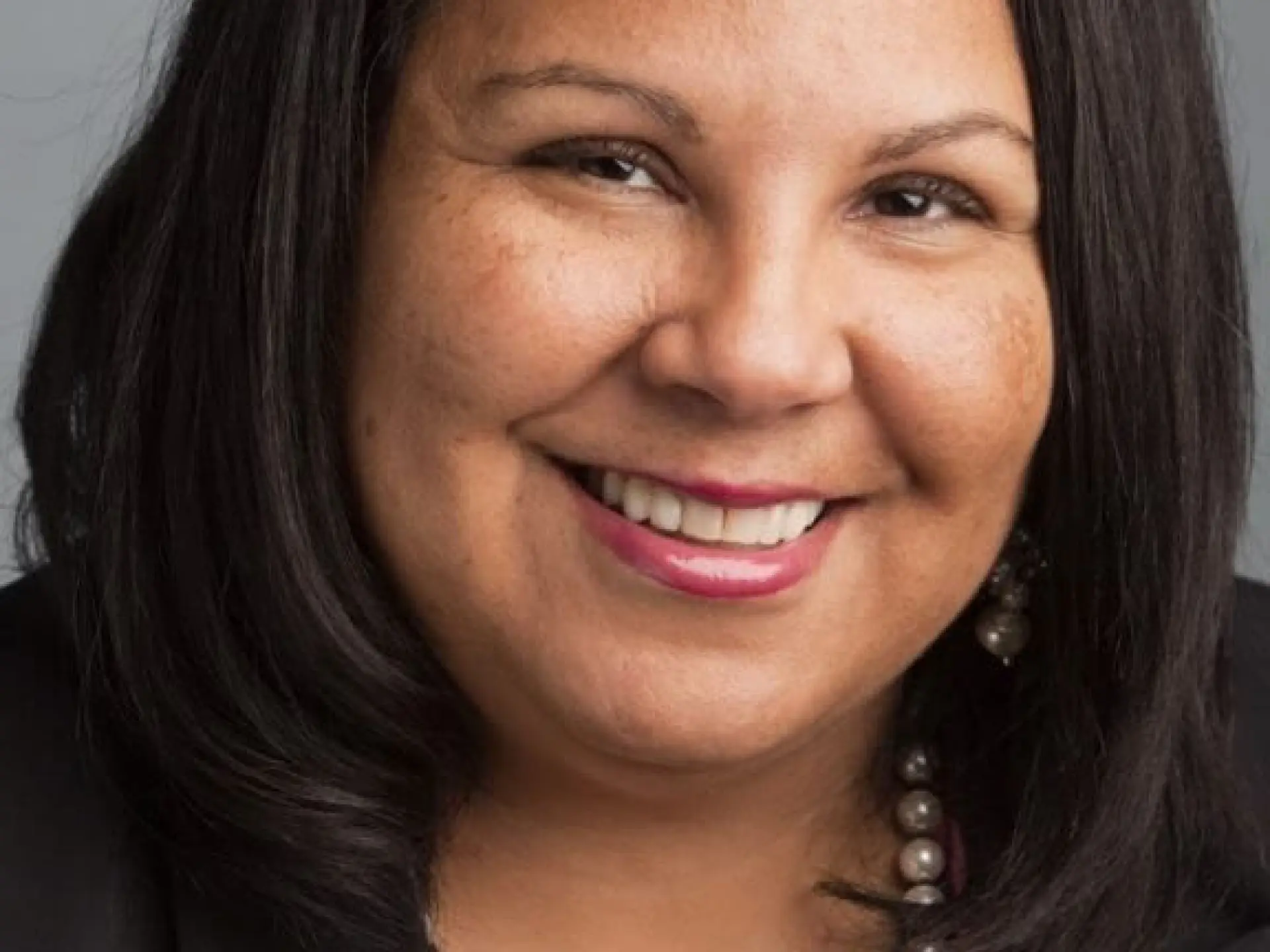 Smiling woman with long dark hair, wearing a black blazer and pearl necklace.