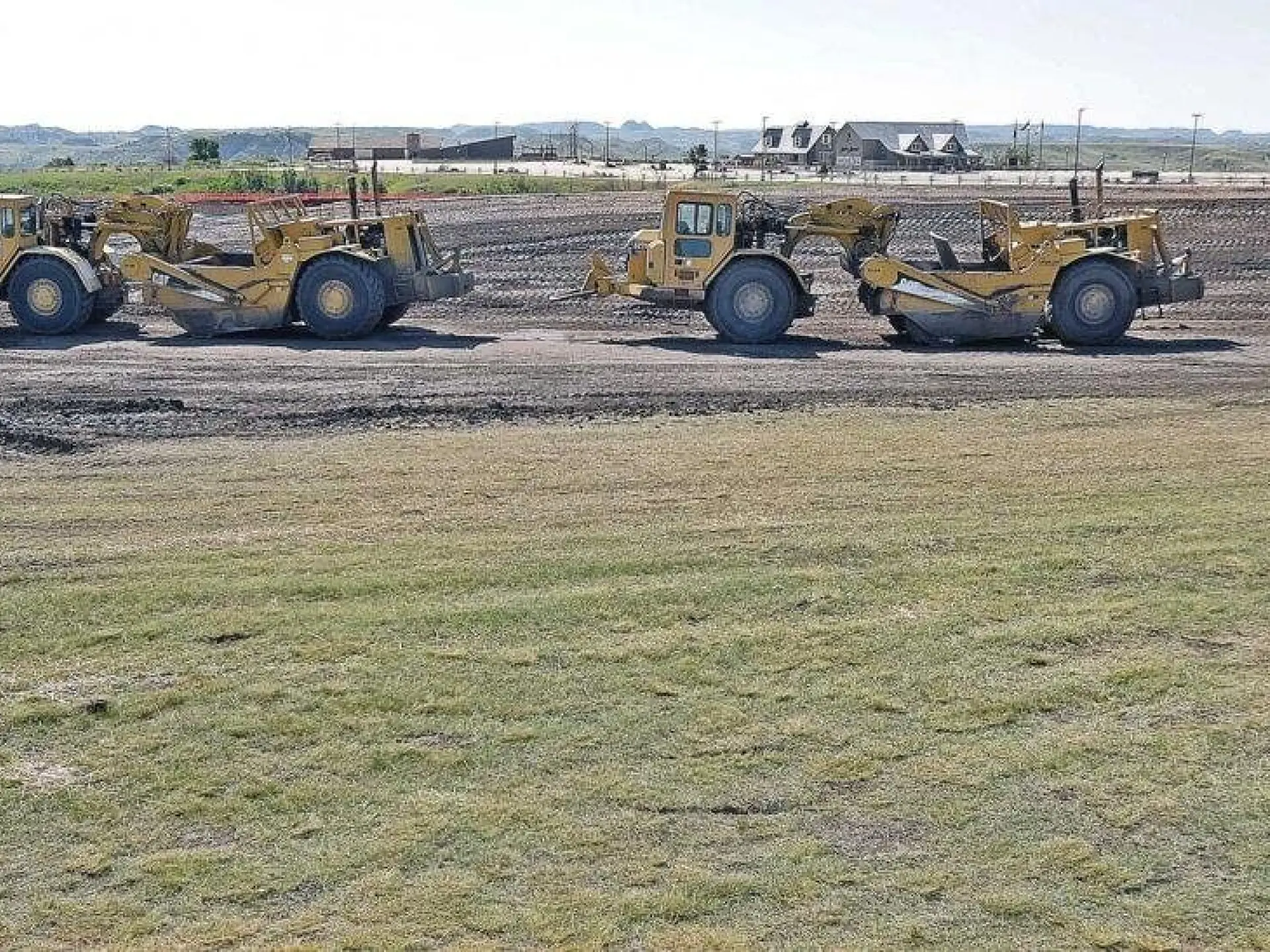 Three yellow construction vehicles on a dirt field with grass in the foreground.