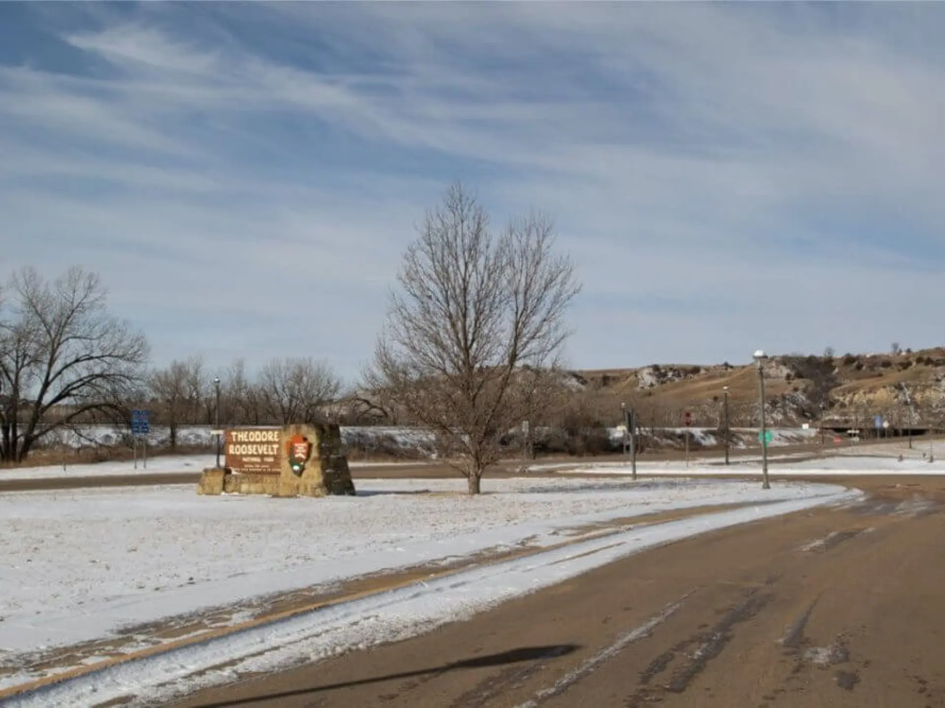 Entrance to park with dry trees, snow-dusted ground, and a clear sky.