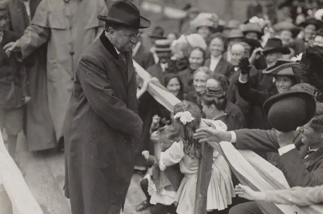 Man in coat and hat cuts ribbon at event, crowd watches eagerly.