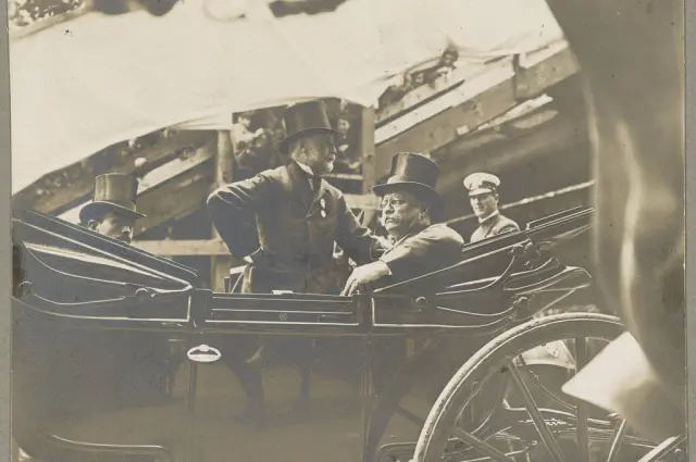 Two men in top hats seated in a horse-drawn carriage, with onlookers around.